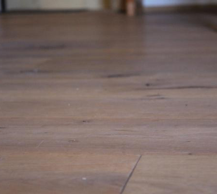 Eine Nahaufnahme eines Holzbodens mit sichtbarer Maserung und leichten Kratzern. Die Beleuchtung ist weich und wirft sanfte Schatten.