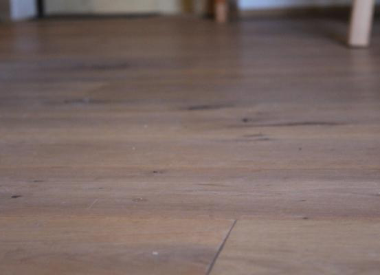 Eine Nahaufnahme eines Holzbodens mit sichtbarer Maserung und leichten Kratzern. Die Beleuchtung ist weich und wirft sanfte Schatten.