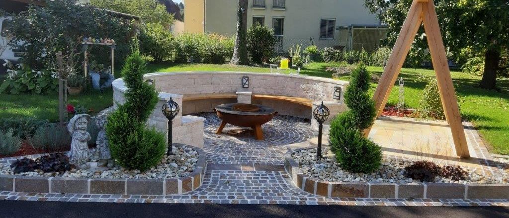Ein Gartenbereich mit Feuerstelle, Bänken und dekorativen Elementen. Bäume, Sträucher und Steine umgeben den Bereich.