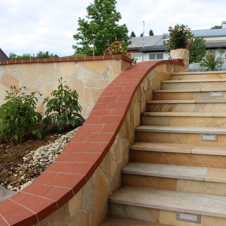 Steinmauer mit einer Treppe und einer gebogenen Ziegelkante, die zu einem Garten mit Pflanzen führt. Bäume und ein Gebäude sind im Hintergrund.