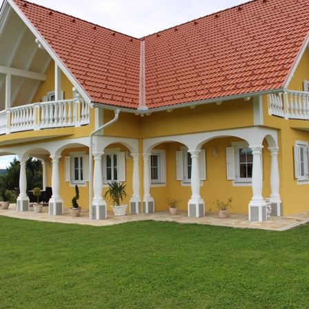 Ein gelbes Haus mit einem roten Dach, weißen Säulen und Balkonen. Es hat mehrere Topfpflanzen und einen großen grünen Rasen davor.