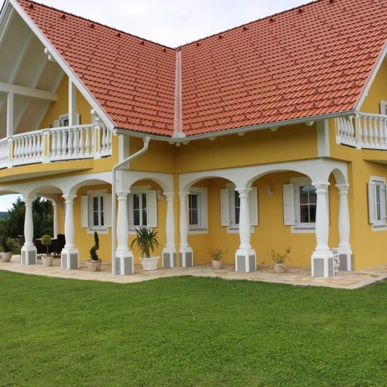 Ein gelbes Haus mit einem roten Dach, weißen Säulen und Balkonen. Es hat mehrere Topfpflanzen und einen großen grünen Rasen davor.