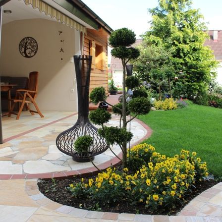 Ein Garten mit einer Terrasse, einem Holzstuhl, einem Tisch und einer dekorativen schwarzen Metallskulptur. Es gibt einen kreisförmigen Garten mit gelben Blumen, einem Hecke und einem Rasen.