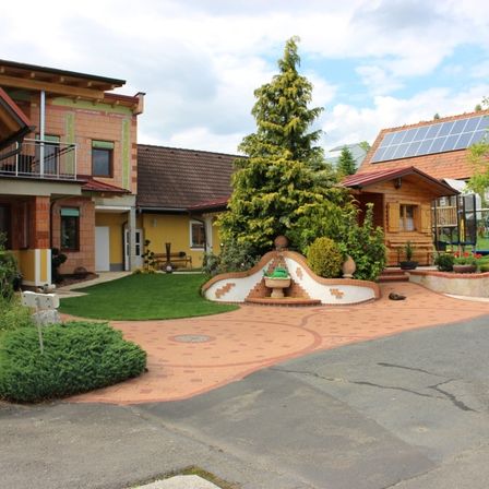 An einem sonnigen Tag zeigt ein Haus mit Ziegelfassade, Solarpanels und einem Garten mit Bäumen, Sträuchern und einem Betonweg.
