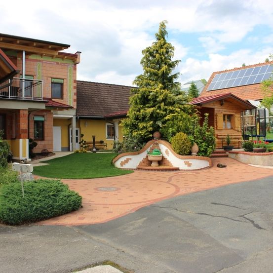 An einem sonnigen Tag zeigt ein Haus mit Ziegelfassade, Solarpanels und einem Garten mit Bäumen, Sträuchern und einem Betonweg.