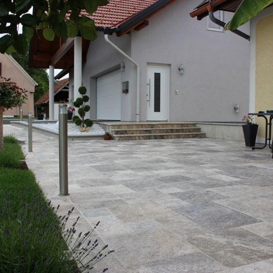 Ein modernes Haus mit einem großen gepflasterten Eingang. Eine Treppe führt zu einer weißen Tür. Blumentöpfe und Büsche schmücken den Eingang.