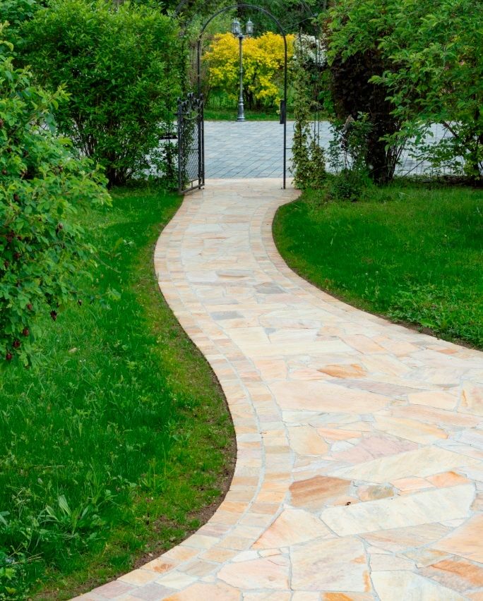 Ein gebogener Steinweg schlängelt sich durch einen üppigen grünen Garten, gesäumt von Büschen und einem schwarzen Tor.