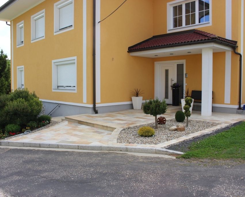 Ein gelbes Haus mit weißen Fenstern hat eine Veranda mit einem braunen Dach. Vor dem Haus stehen Topfpflanzen und Sträucher. Eine Einfahrt führt zum Eingang.