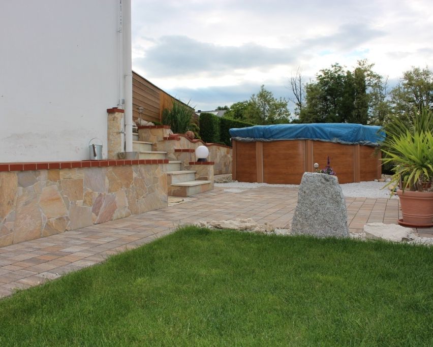 Ein Garten mit Steinmauer, Treppe, hölzerne Whirlpool und überdachter Terrasse. Ein Topf mit Pflanze und Lampe sind in der Nähe.