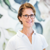 Eine Frau mit Brille und einem weißen Polohemd steht lächelnd vor einem Hintergrund aus grünen Blättern.