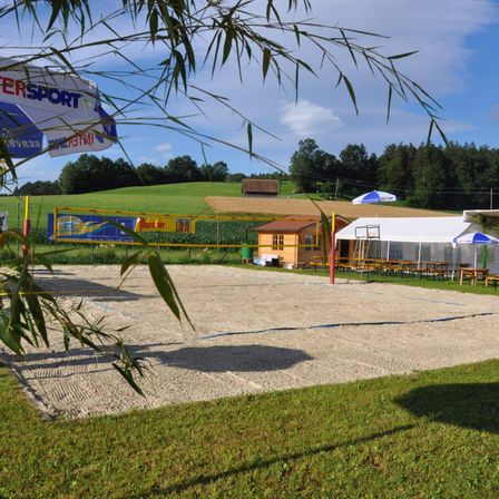 Ein Volleyballfeld umgeben von Gras mit Blick aufs Land. Ein kleines Haus und ein weißes Zelt sind in der Nähe.