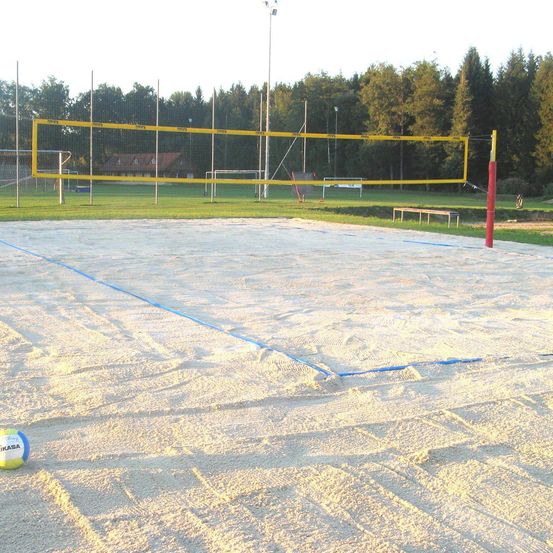 Ein Outdoor-Volleyballfeld ist mit Sand bedeckt. Das Netz ist gelb mit blauen Linien auf dem Sand. In der Ferne gibt es ein Gebäude und ein Fußballtor.