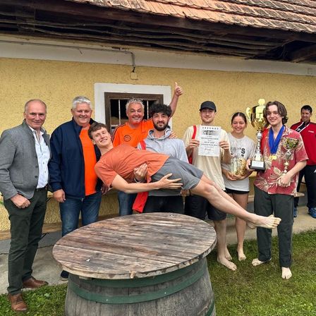 Gruppe von Menschen in ländlicher Umgebung, posiert für ein Foto mit Trophäen und Urkunden. Ein Mann liegt mit erhobenen Füßen auf einem Fass.