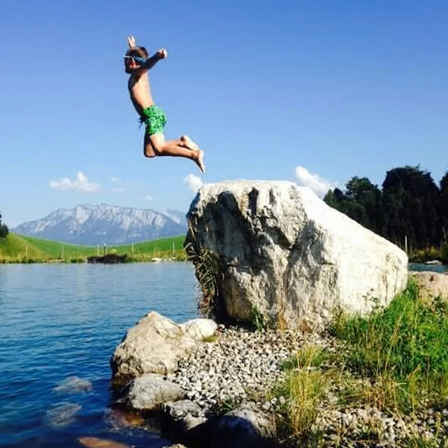 Ein junger Junge springt von einem großen Felsen in den See. Das Wasser ist ruhig, und im Hintergrund befinden sich Berge und ein klarer blauer Himmel.