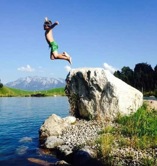 Ein junger Junge springt von einem großen Felsen in den See. Das Wasser ist ruhig, und im Hintergrund befinden sich Berge und ein klarer blauer Himmel.