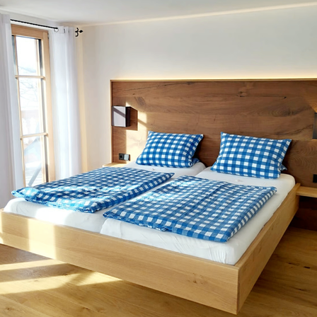 Ein Schlafzimmer mit einem Holzbett, zwei blau karierten Betten und einem Holzkopfteil. Der Raum hat weiße Wände, ein Glasfenster mit Vorhängen und Holzböden.
