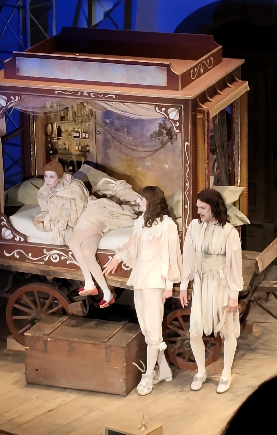 Drei Darsteller auf einer Bühne, einer sitzt auf einem Bett, die anderen beiden stehen davor. Das Bett ist mit Kissen dekoriert und hat eine Wand mit einem Gemälde dahinter.