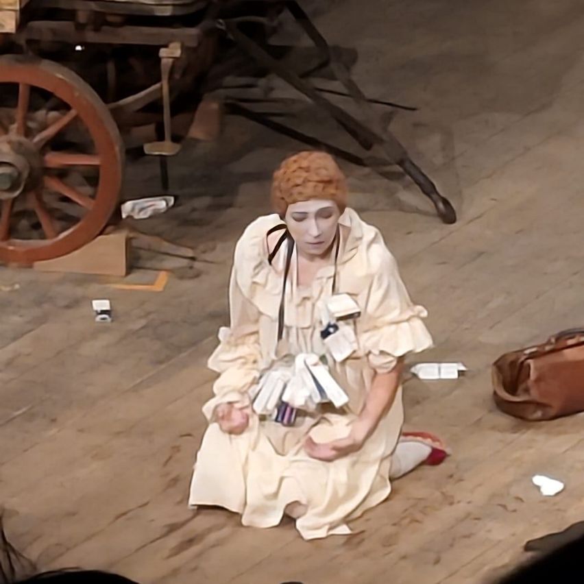 Eine Frau in einem Vintage-Kleid sitzt mit überkreuzten Beinen auf einem Holzboden und hält eine Sammlung von Gegenständen. Ein Rad und eine braune Tasche sind in der Nähe. Der Boden ist schmutzig, und links steht ein Karton.