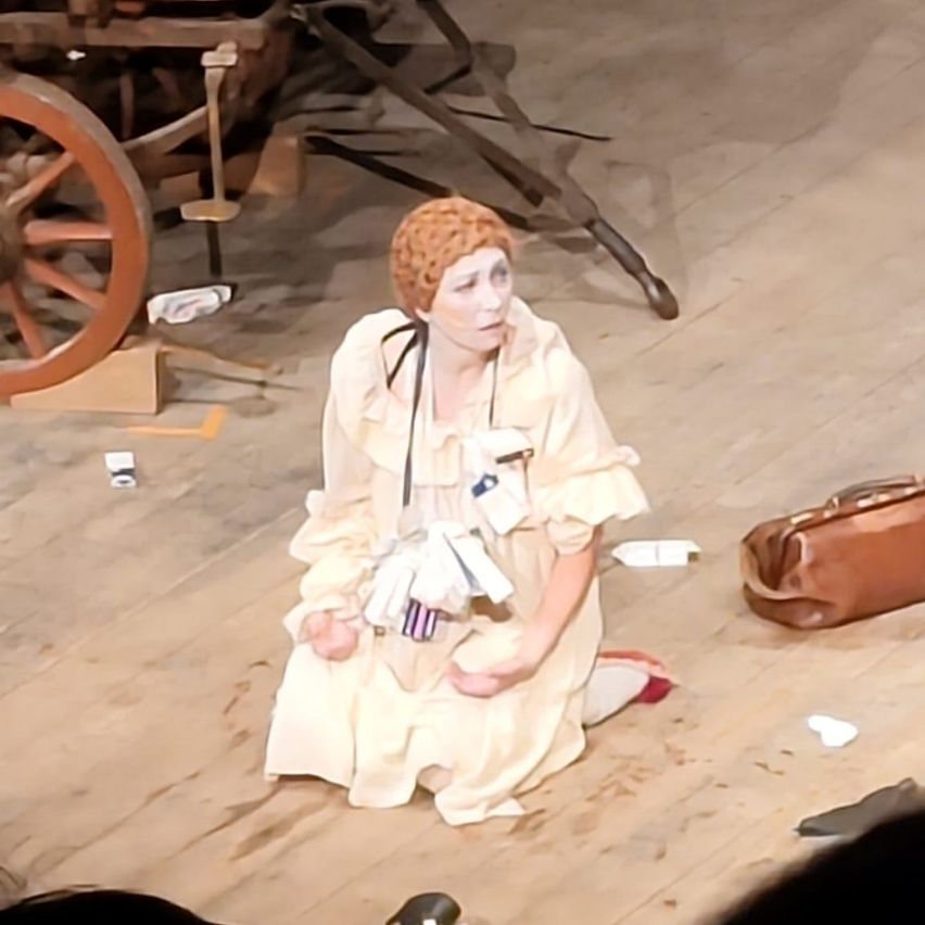 Eine Frau in einem weißen Kleid kniet auf einem Holzboden, möglicherweise auf einer Bühne. Sie hält Papiere und trägt eine schwarze Schleife. Ein brauner Beutel ist in der Nähe und ein großes Rad ist hinter ihr.