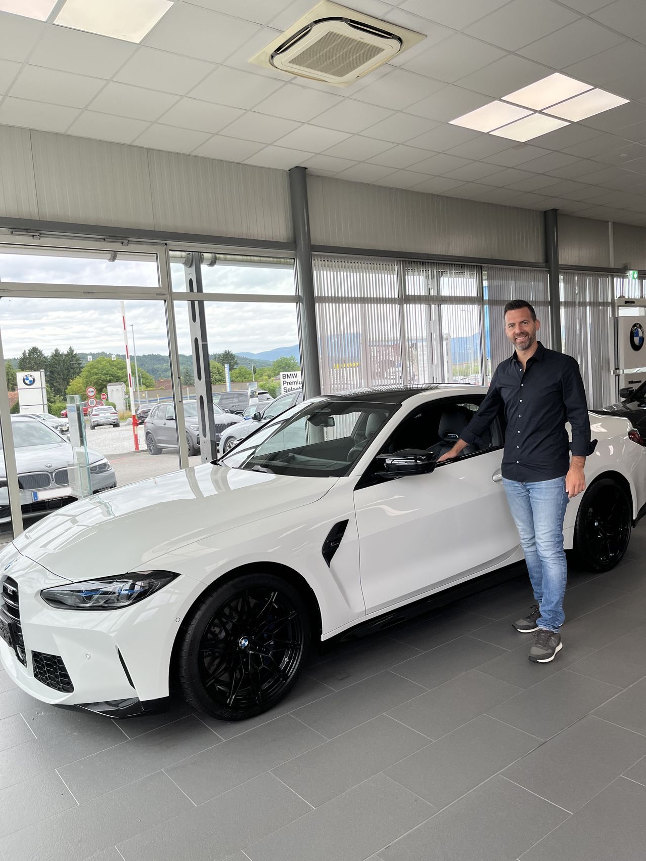 Ein Mann steht neben einem weißen BMW M4 Coupé in einer Ausstellung mit Glaswänden, umgeben von anderen Autos.