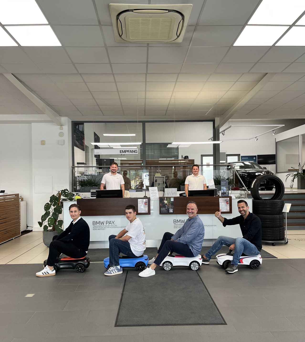 Eine Gruppe von Menschen sitzt auf Spielzeugautos in einem BMW-Showroom. Vier Männer sitzen auf den Autos, während zwei hinter der Theke stehen.