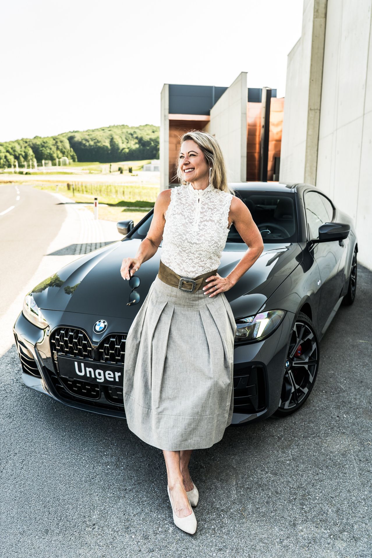 Eine Frau steht neben einem schwarzen BMW M240i Auto, hält eine Sonnenbrille und lächelt. Das Auto hat ein Nummernschild mit dem Wort Unger.