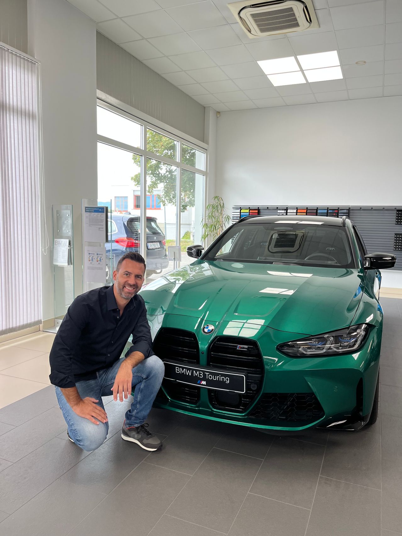 Ein Mann hockt neben einem grünen BMW M3 Touring in einem Showroom und präsentiert dessen schlankes Design und moderne Funktionen.