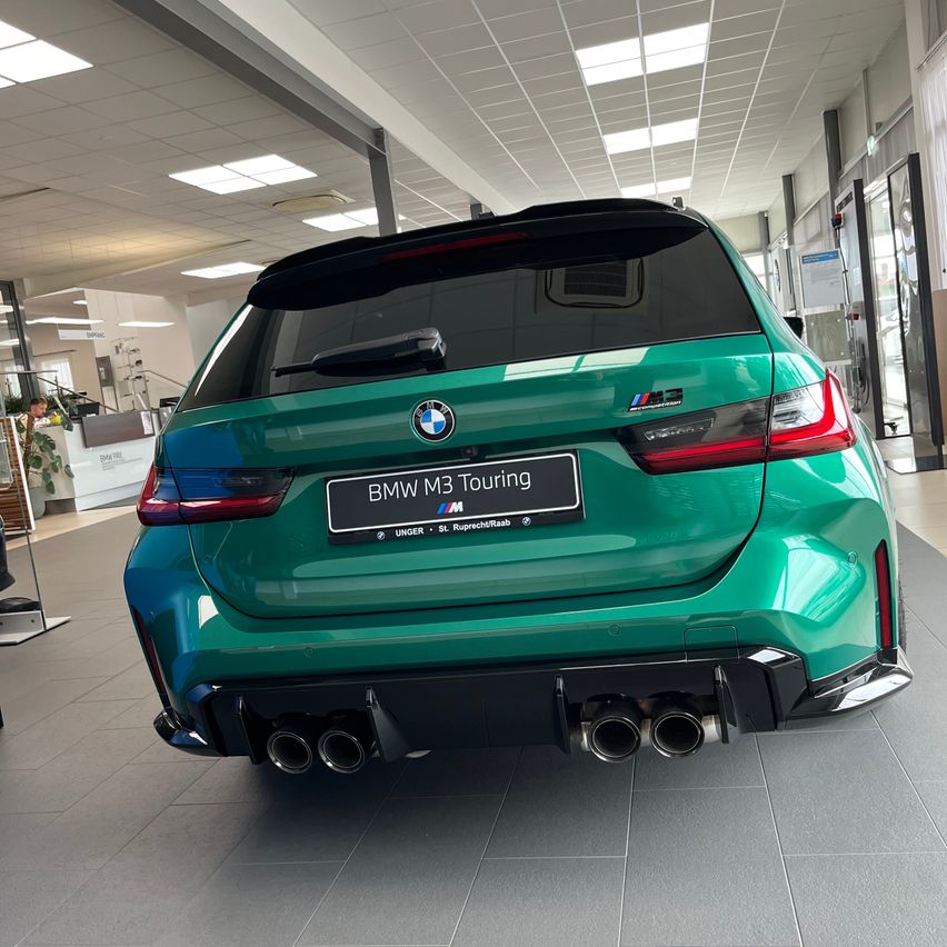 Die Rückseite eines grünen BMW M3 Touring ist in einer Ausstellung mit grauen Fliesenböden geparkt. Das Auto hat vier Auspuffrohre und ein Nummernschild, auf dem BMW M3 Touring steht.