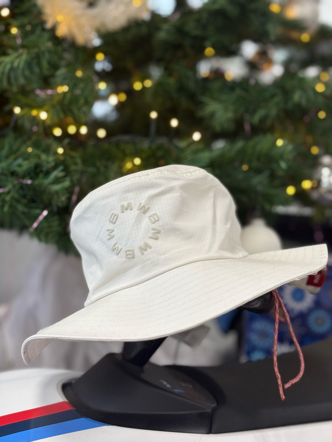 Ein weißer Bucket-Hut mit '3MW8M3W' im kreisförmigen Text wird vor einem beleuchteten Weihnachtsbaum gezeigt.
