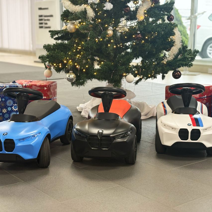 Drei Spielzeugautos in Blau, Schwarz und Weiß sind in einer Reihe unter einem geschmückten Weihnachtsbaum geparkt. Hinter dem Baum ist ein Auto durch ein Glasfenster sichtbar.