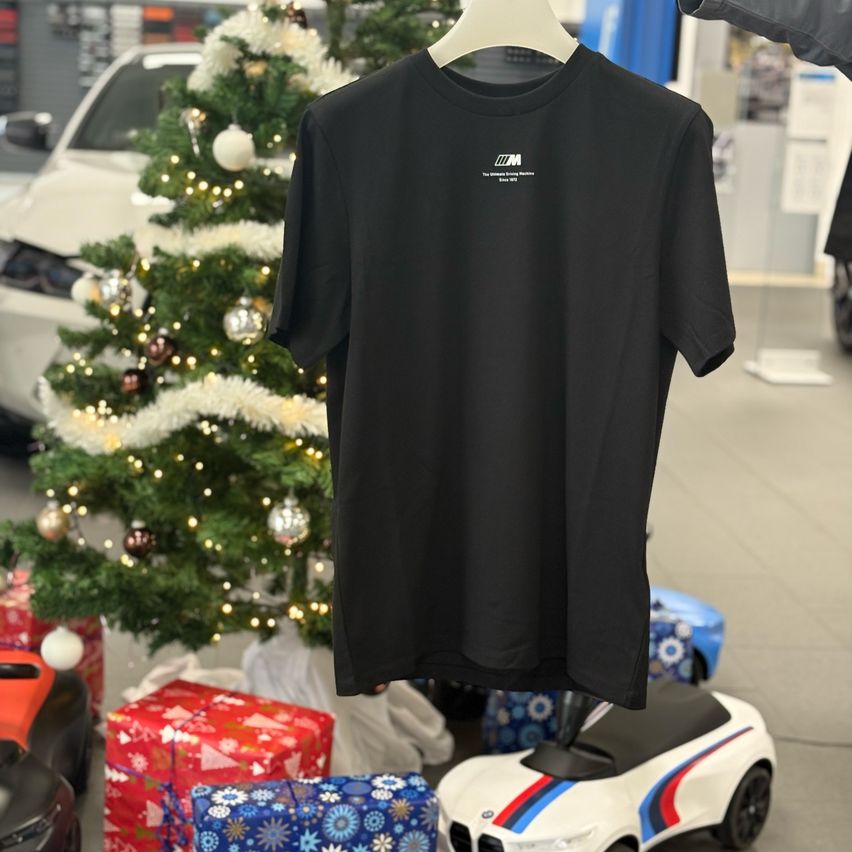 Ein schwarzes T-Shirt hängt an einem Kleiderbügel vor einem festlichen Hintergrund mit einem Weihnachtsbaum, Geschenken und einem weißen Auto. Das T-Shirt hat ein weißes Logo.