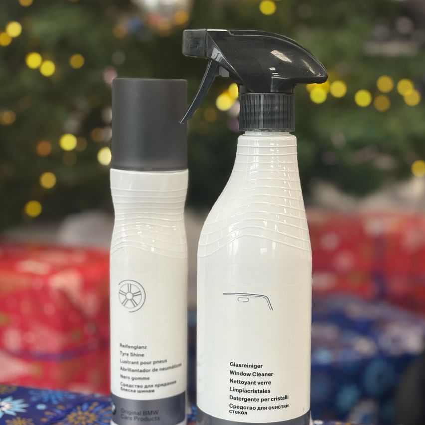 Zwei weiße Sprühflaschen mit schwarzen Kappen auf einer blauen Oberfläche. Ein Weihnachtsbaum ist im Hintergrund. Die linke Flasche ist mit 'Reifenglanz Reifenglanz' und die rechte Flasche mit 'Glaschreiniger Window Cleaner' beschriftet.