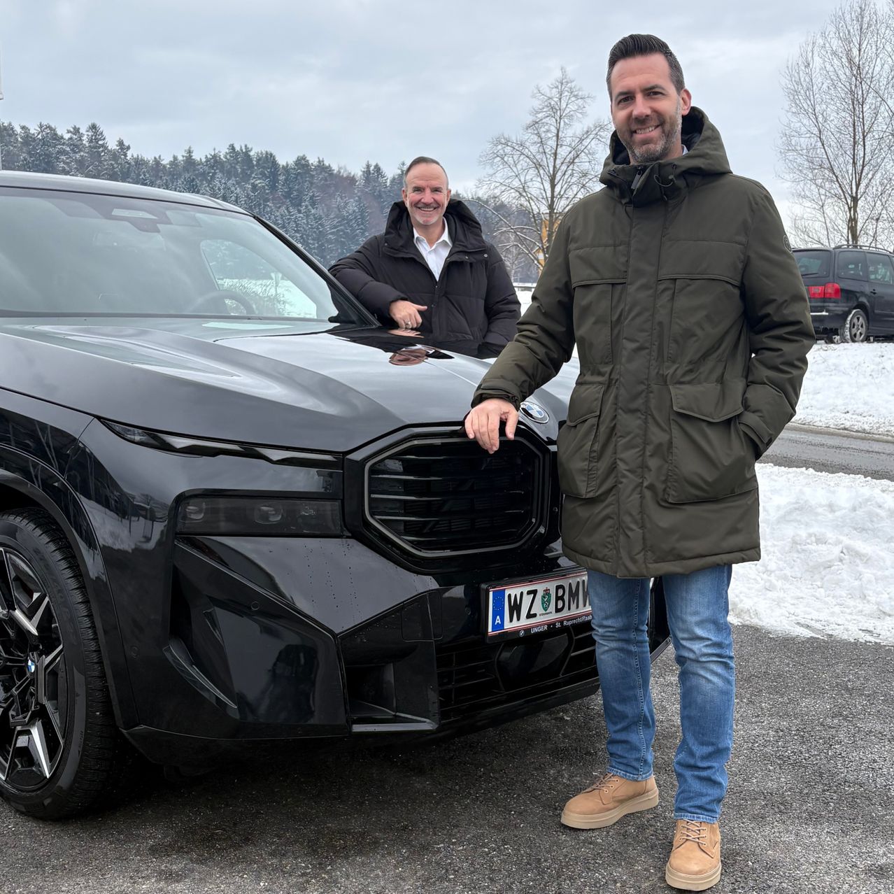 Zwei Männer stehen lächelnd neben einem schwarzen BMW-Fahrzeug, mit einem verschneiten Hintergrund.
