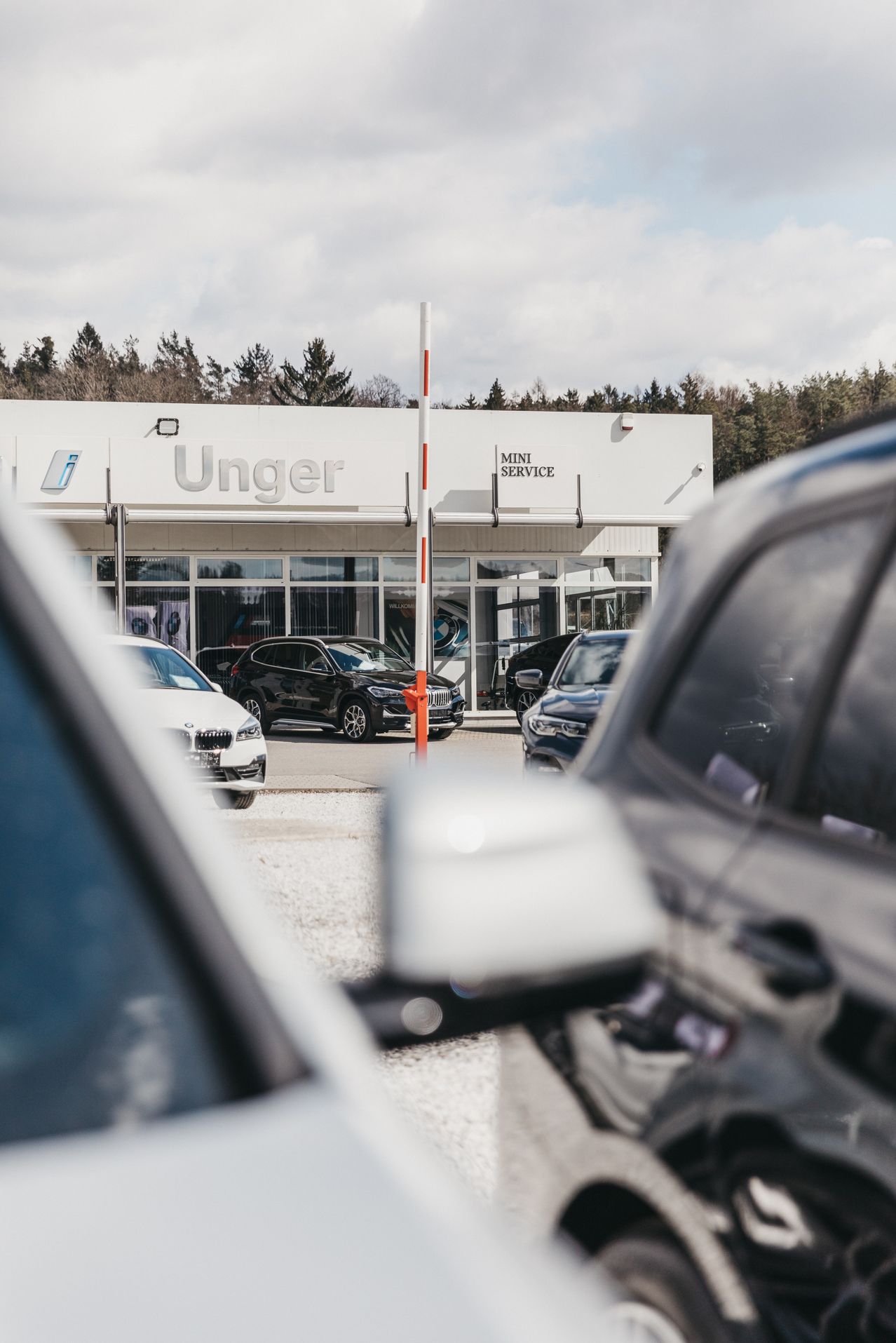 Ein BMW-Händler namens Unger wird mit mehreren geparkten Autos davor gezeigt. Das Gebäude hat große Fenster und ein MINI SERVICE-Schild. Bäume sind im Hintergrund sichtbar.