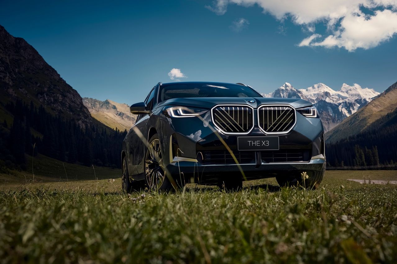 The front view of a black BMW X3 SUV is parked in a grassy field with mountains in the background.