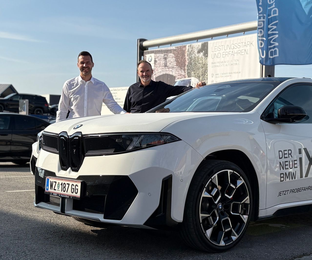 Zwei Männer posieren neben einem weißen BMW SUV. Der Mann auf der linken Seite lächelt, während der Mann auf der rechten Seite sich auf das Auto stützt. Das Nummernschild des Autos lautet 187 GG.