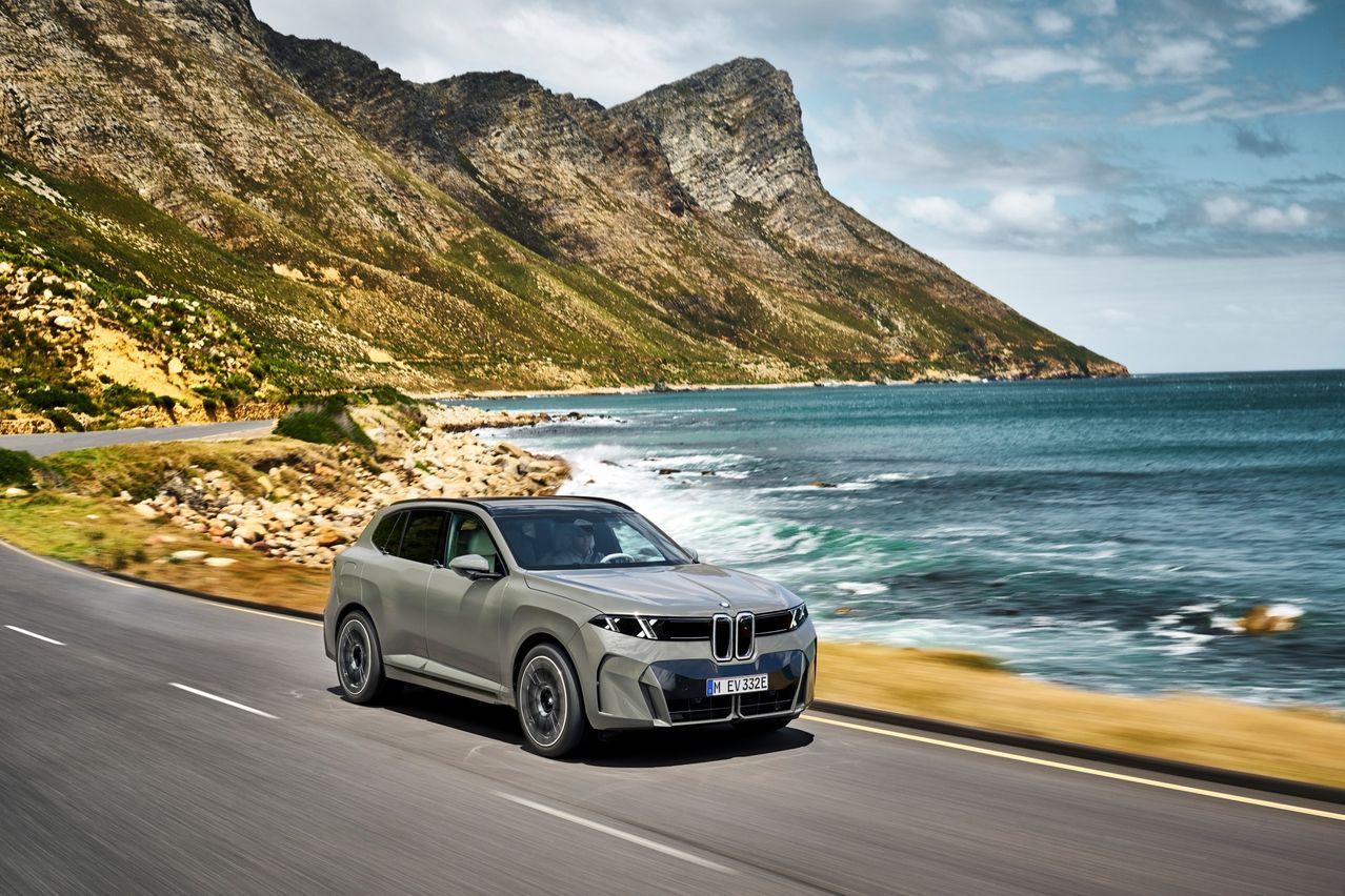 A gray BMW SUV drives along a coastal road with mountains and ocean in the background.