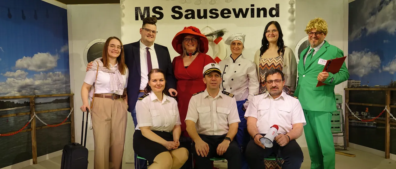 Eine Gruppe von Menschen in Uniform und Hüten posiert für ein Foto auf einer Bühne mit einem Schild, das MS Sausewind liest.