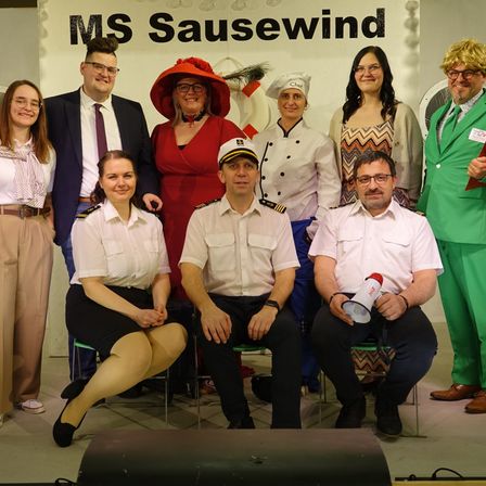 Eine Gruppe von Menschen in Kostümen, wahrscheinlich für eine Aufführung, steht und sitzt vor einer Wand mit dem Text 'MS Sausewind'.
