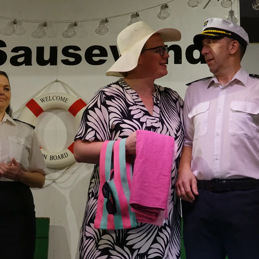Drei Menschen stehen vor einer Wand mit dem Text 'Sausev' und einem Rettungsring. Eine Frau in Hut und Brille hält ein rosa Handtuch und eine Tasche. Ein Matrose und eine weitere Frau schauen zu ihr.