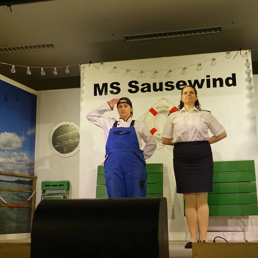 Zwei Personen in Kostümen stehen auf der Bühne, einer in einer Latzhose, der andere in einer Uniform. Sie stehen einem Publikum gegenüber. Ein Rettungsring hängt hinter ihnen am Hintergrund.