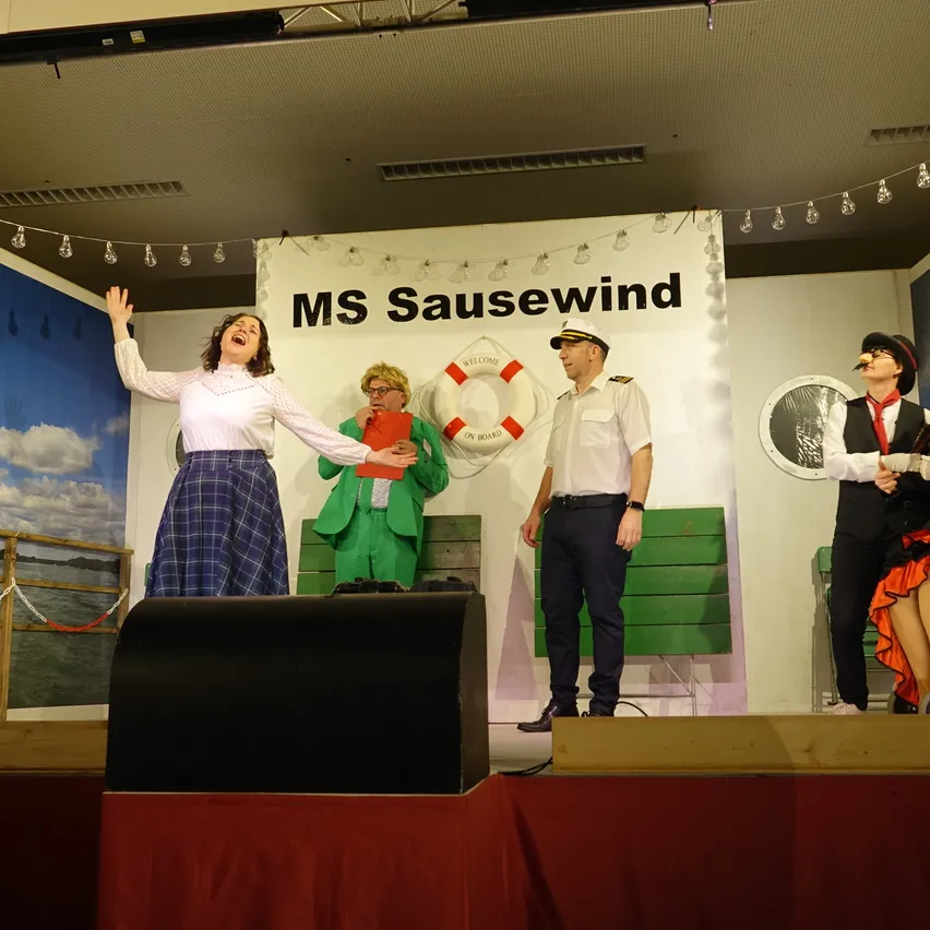Eine Gruppe von Menschen auf der Bühne, eine Frau lacht, während eine andere einen roten Gegenstand hält. Hinter ihnen steht ein Schild mit MS Sausewind und einem Rettungsring.