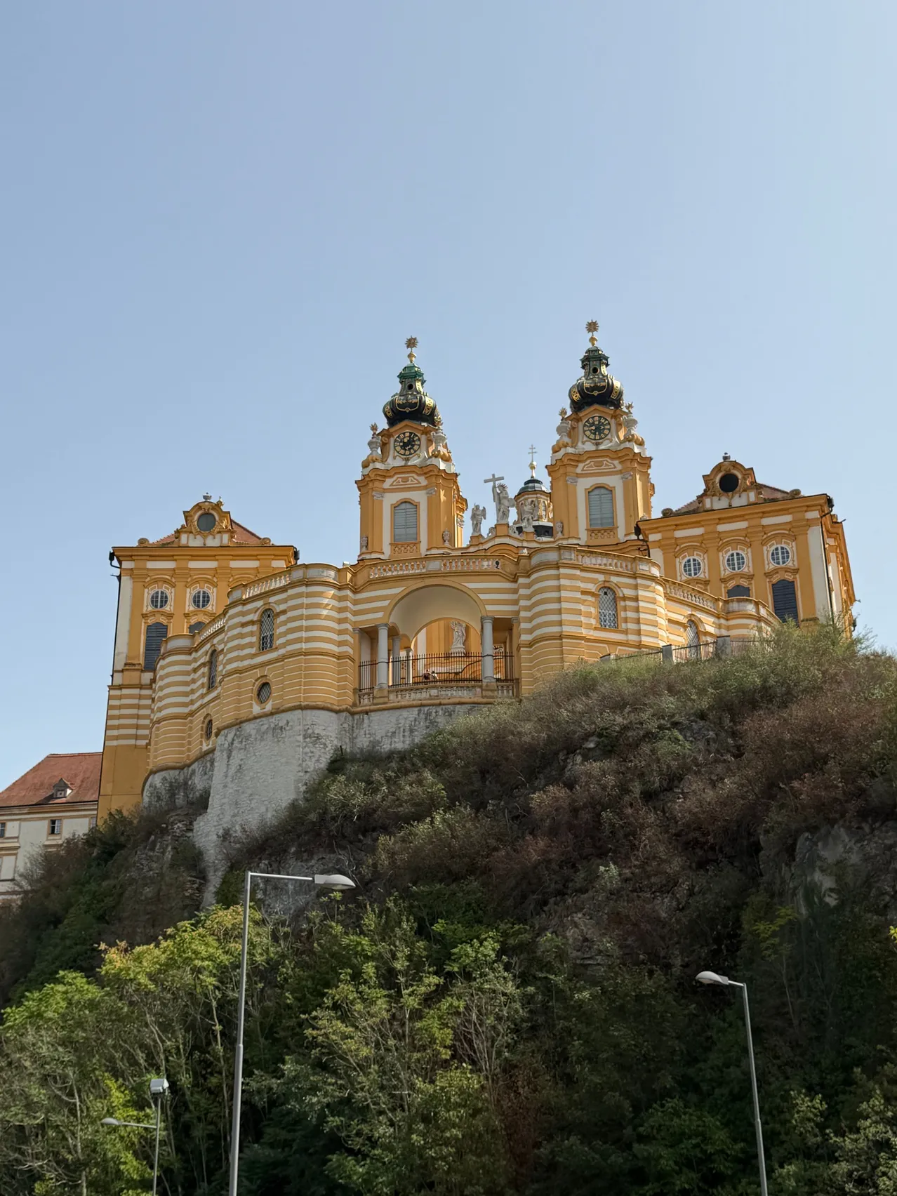 Eine große gelbe und weiße Barockkirche mit zwei Türmen und einer Uhr auf der Spitze steht auf einem Hügel unter einem klaren blauen Himmel.