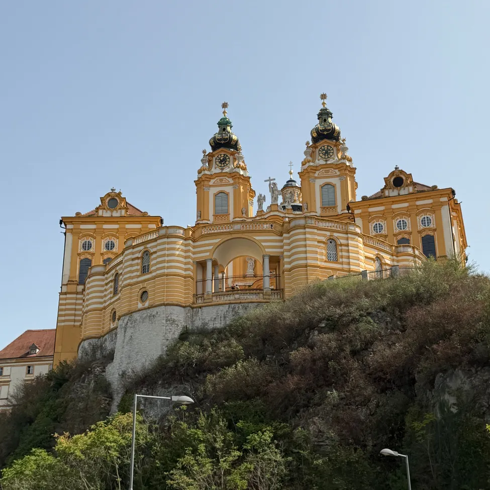 Eine große gelbe und weiße Barockkirche mit zwei Türmen und einer Uhr auf der Spitze steht auf einem Hügel unter einem klaren blauen Himmel.