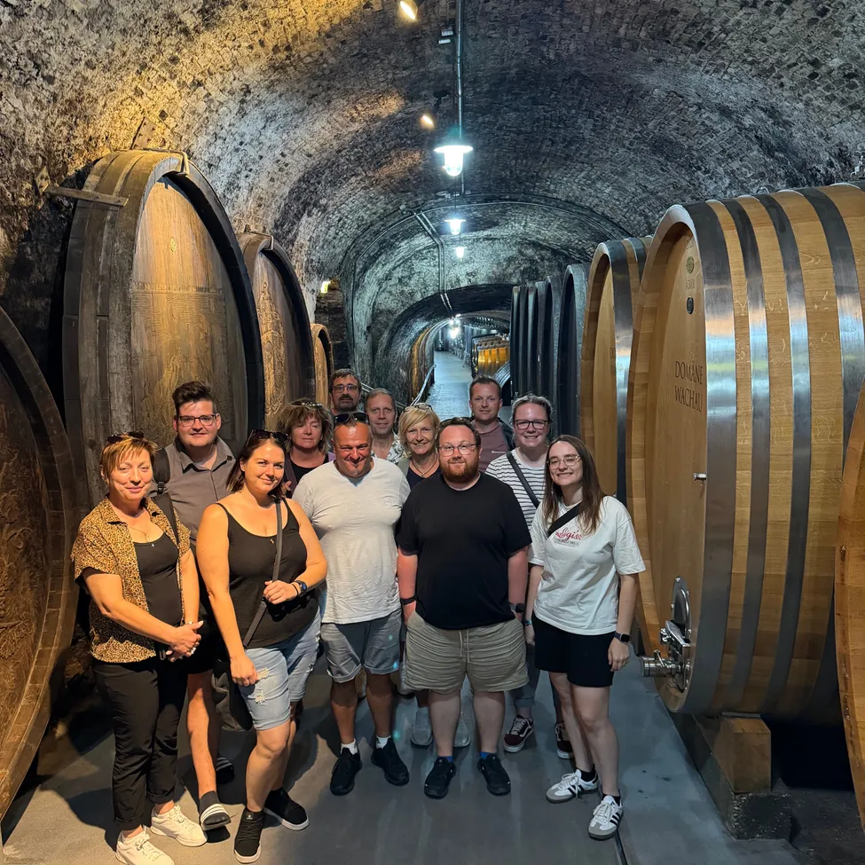 Eine Gruppe von Menschen posiert für ein Foto in einem Weinkeller mit großen Holzfässern.