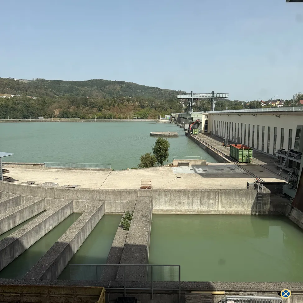 Ein großes Reservoir mit einem Betondamm, umgeben von einer Betonmauer und einem Gebäude. Bäume und Berge sind im Hintergrund.