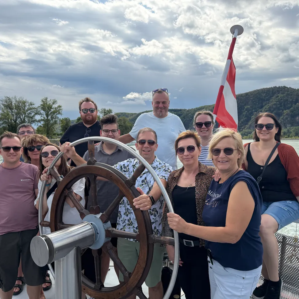 Eine Gruppe von Menschen auf einem Boot, posiert für ein Foto mit einem Lenkrad im Vordergrund. Dahinter eine Flagge und eine malerische Aussicht auf Berge und Bäume.