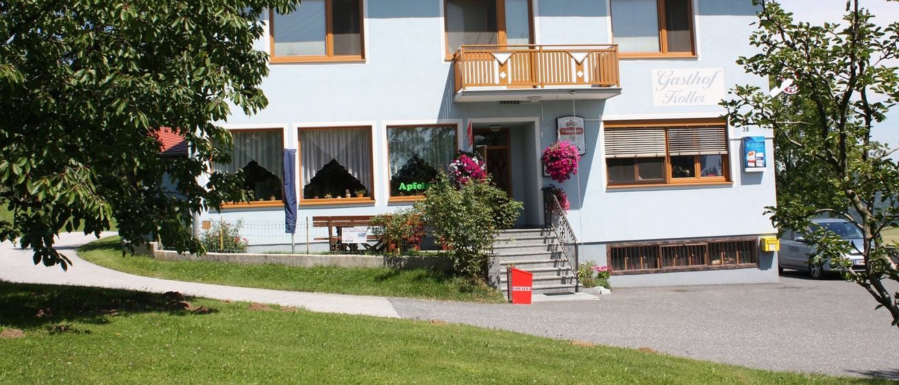Ein hellblaues Haus mit Solarpanelen auf dem Dach, einem Balkon mit Holzgeländer und einem Schild, das Gasthof Koller anzeigt. Das Haus hat mehrere Fenster, einige mit weißen Vorhängen. Ein roter Mülleimer steht vor dem Haus.