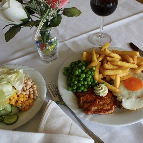 Ein weißer Tischläufer mit zwei Tellern mit Essen, einer Blumenvase und einem Weinglas. Ein Teller enthält Pommes Frites, ein Spiegelei und ein Gemüsegericht. Der andere Teller hat Salat, Bohnen, Mais und Gurken.
