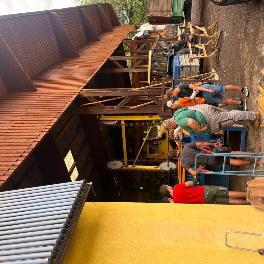 Eine Gruppe von Männern arbeitet auf einer Baustelle. Sie stehen um eine gelbe Maschine mit einem Rad und einer Metallstruktur herum. Sie tragen Hüte und Turnschuhe. Das Gebiet ist von Holzstrukturen und einer gelben Wand umgeben.